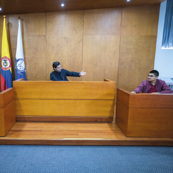 Escenario de simulación judicial con dos personas en estrados opuestos dentro de una sala de audiencias, con banderas oficiales al fondo y paredes de madera.