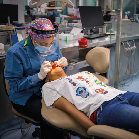 Una persona con uniforme y guantes realiza un procedimiento dental a un paciente recostado en una silla odontológica dentro de un consultorio equipado.