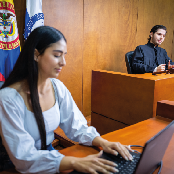 Escenario de simulación judicial con una persona escribiendo en un computador portátil en primer plano y otra persona en el estrado, detrás de un escritorio, con banderas oficiales al fondo.