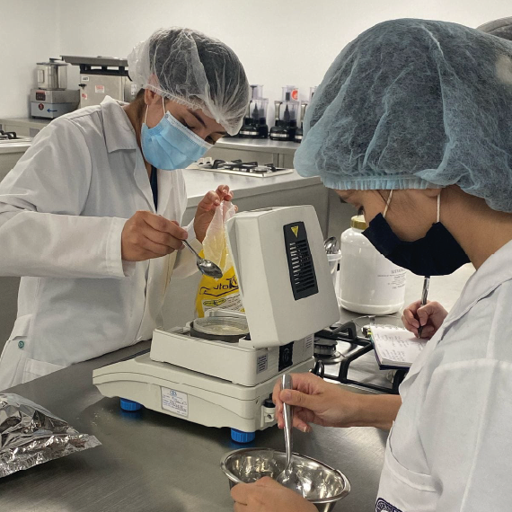 Personas en laboratorio pesan ingredientes en una balanza digital y utilizan utensilios metálicos, usando batas y gorros de protección.