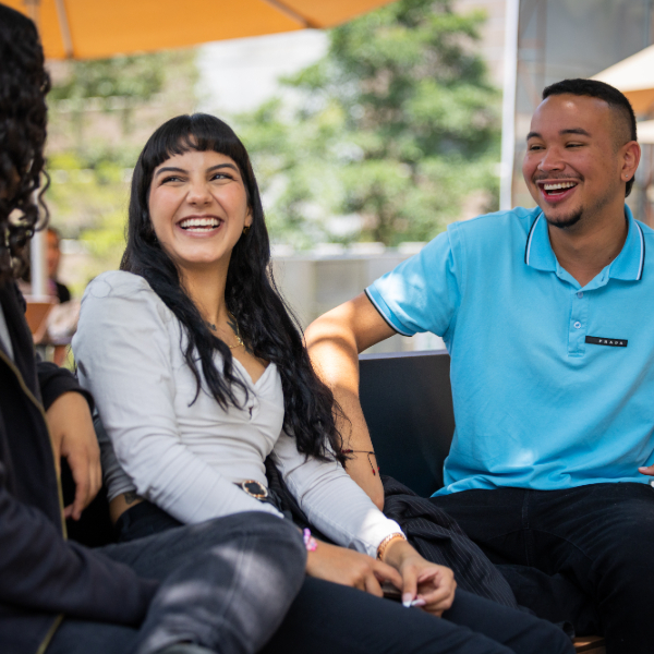 Tres personas están sentadas al aire libre, conversando en un espacio con vegetación y sombrillas en el fondo.