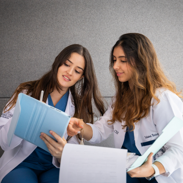 Dos personas con batas blancas revisando documentos y escribiendo en una tableta en un entorno educativo.