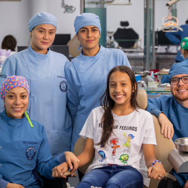 Varias personas con uniforme odontológico están junto a una persona sentada en una silla dental, en un consultorio equipado para prácticas clínicas.