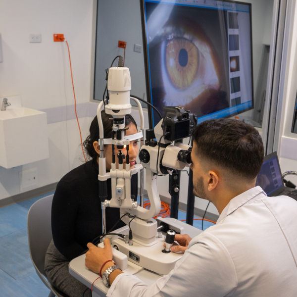 Profesional de la salud realiza examen ocular con una lámpara de hendidura a una paciente, mientras una imagen ampliada del ojo se proyecta en una pantalla al fondo.