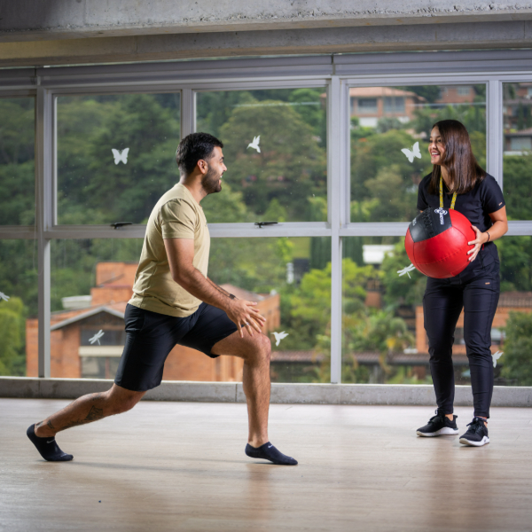 Dos personas en un salón con ventanales; una realiza una estocada mientras la otra sostiene un balón medicinal.
