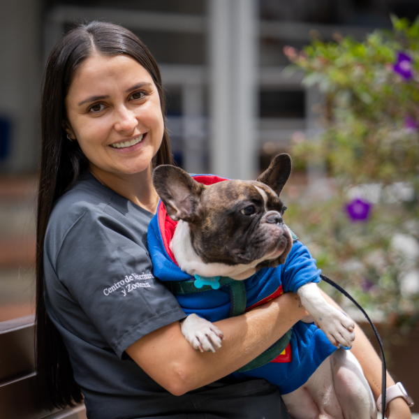 Persona sostiene en brazos a un perro con ropa colorida, en un espacio exterior con plantas al fondo.