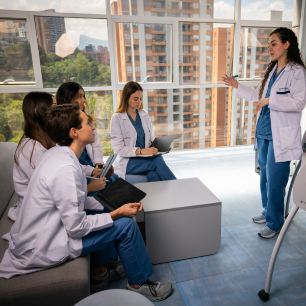 Grupo de personas con batas blancas y uniformes clínicos participa en una sesión académica, mientras una persona expone frente a ellas en un aula con ventanales y vista a edificios