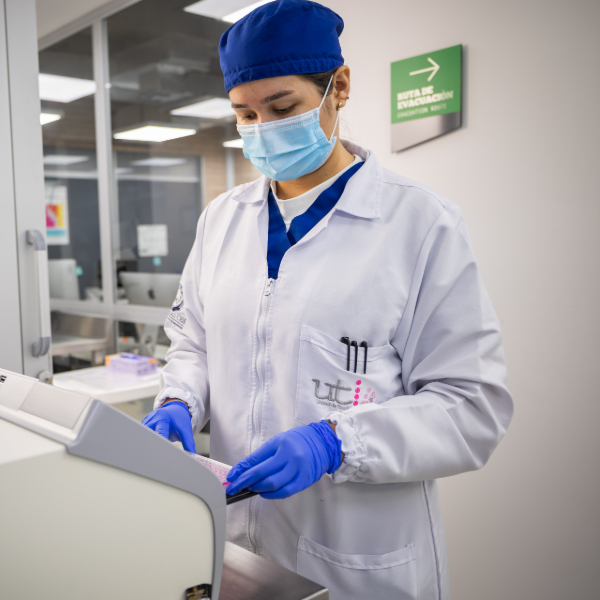 Persona con bata blanca, gorro azul y guantes azules manipula un equipo médico o de laboratorio en un entorno clínico. Al fondo se observan paredes claras, señalización verde y mobiliario propio de una instalación sanitaria.