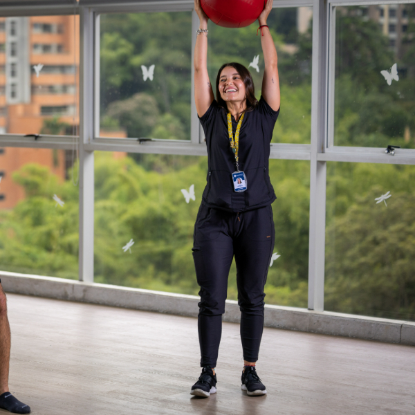 Persona de pie en un espacio interior con ventanales, sosteniendo una pelota roja sobre la cabeza. Viste ropa deportiva oscura y lleva un gafete colgado al cuello. Al fondo se observa vegetación y edificios a través de las ventanas.