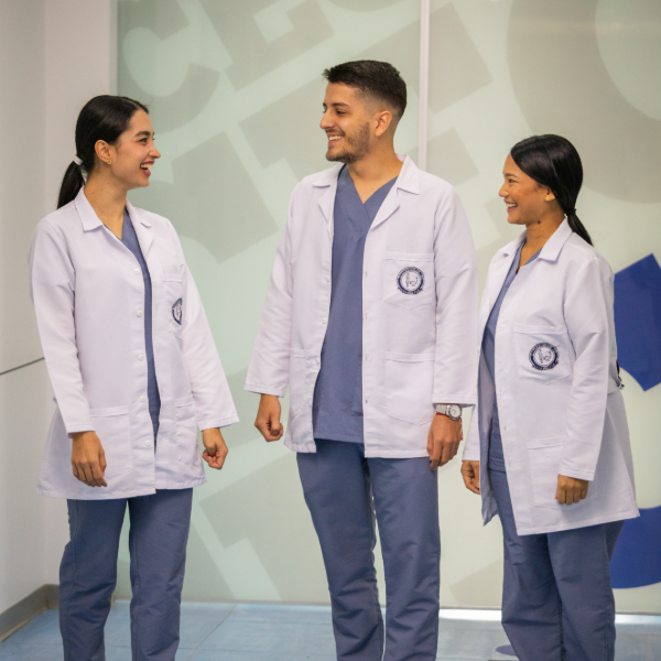 Tres personas con uniformes médicos y batas blancas conversan en un pasillo interior. Cada una lleva ropa clínica azul debajo de la bata y el entorno es limpio y bien iluminado, típico de una instalación hospitalaria o académica.