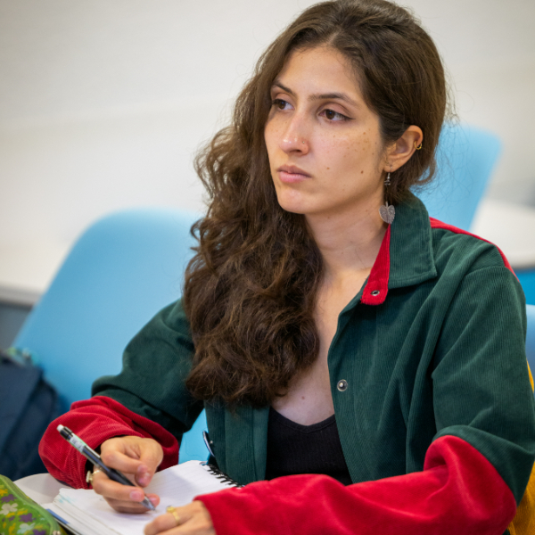 Persona sentada en un aula o sala, escribiendo en una libreta con bolígrafo. Viste chaqueta verde con detalles rojos y hay sillas azules en el fondo.