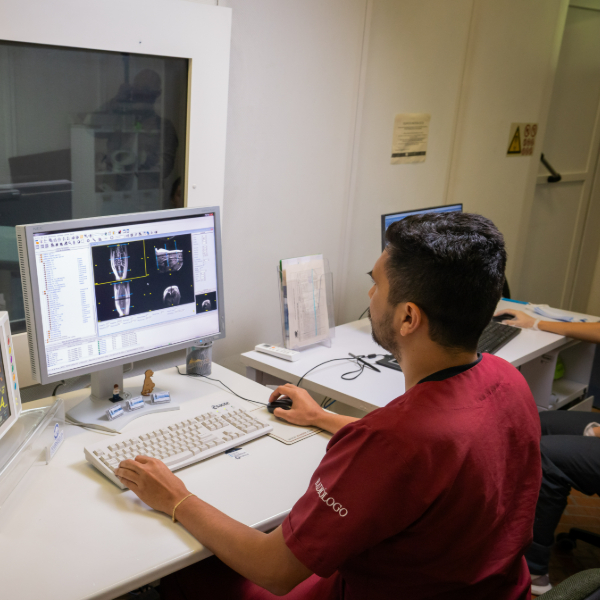 Persona sentada frente a un escritorio utilizando una computadora que muestra imágenes médicas en pantalla, posiblemente radiografías o tomografías. En la mesa hay teclado, ratón y otros elementos de trabajo en un entorno clínico.
