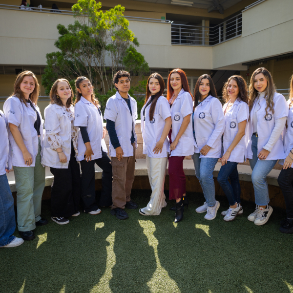 Grupo de personas con batas blancas y uniformes clínicos posan de pie en un espacio exterior con césped, frente a un edificio con balcones y árboles