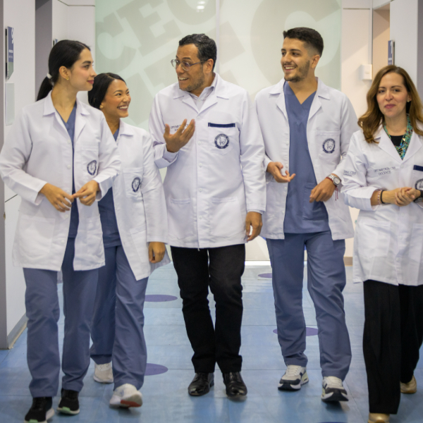 Cinco personas con batas blancas caminan por un pasillo, conversando entre sí.