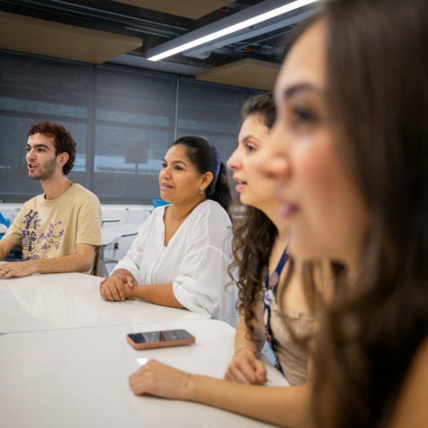 Varias personas están sentadas alrededor de una mesa blanca en un aula, con un teléfono móvil sobre la mesa.