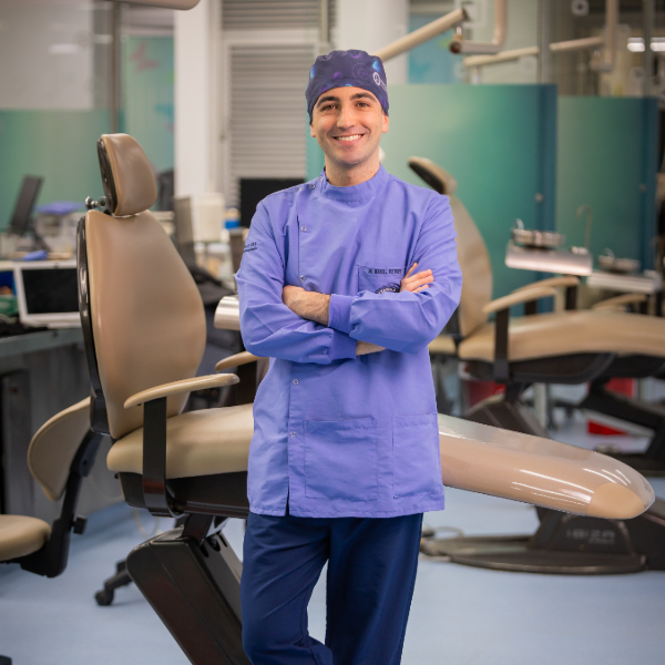 Persona con uniforme clínico azul está de pie con los brazos cruzados en un consultorio odontológico, junto a una silla dental y equipos en el fondo.