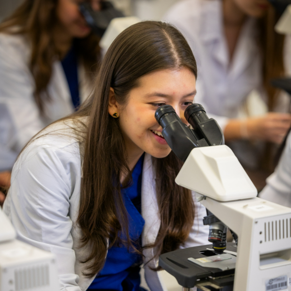 Persona con bata blanca observando a través de un microscopio en un entorno de laboratorio, con otros microscopios y personas al fondo.