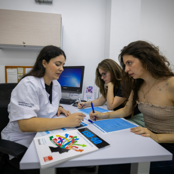 Tres personas están sentadas alrededor de una mesa en un entorno educativo, trabajando con hojas impresas y bolígrafos. Sobre la mesa hay un cuaderno con diseño colorido y una computadora en el fondo.