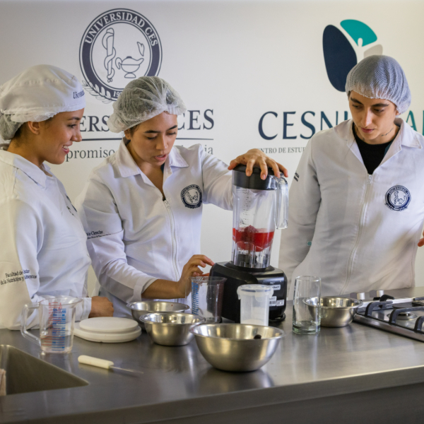 Tres personas con uniformes blancos y gorros de cocina están en una mesa de trabajo en un laboratorio gastronómico, utilizando una licuadora con frutas y rodeadas de recipientes metálicos y utensilios de cocina. En la pared se observan logotipos institucionales.