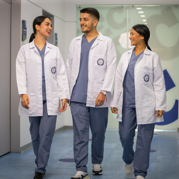 Tres personas caminan por un pasillo, vestidas con uniformes médicos y batas blancas, llevando el emblema institucional en la parte superior izquierda de la bata.