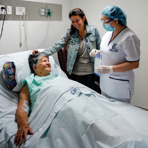 Una persona está recostada en una cama hospitalaria con una vía intravenosa en el brazo, acompañada por otra persona al lado y un profesional de la salud con uniforme y guantes sosteniendo material médico.