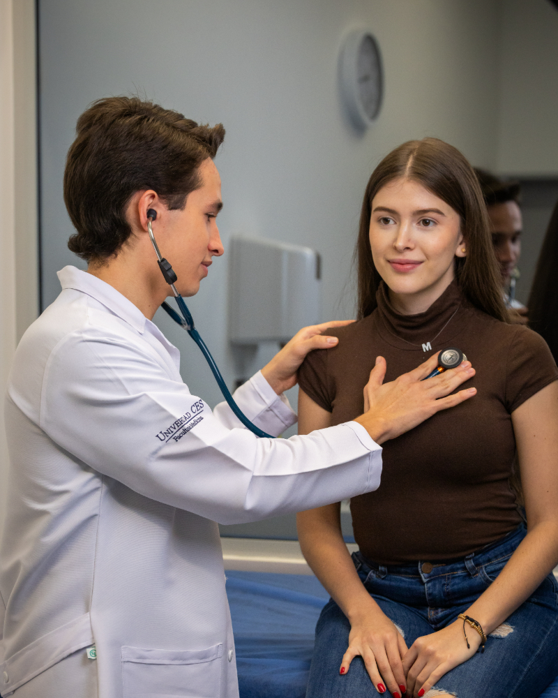 Profesional de la salud utilizando un estetoscopio para auscultar el pecho de una persona sentada en una consulta médica.