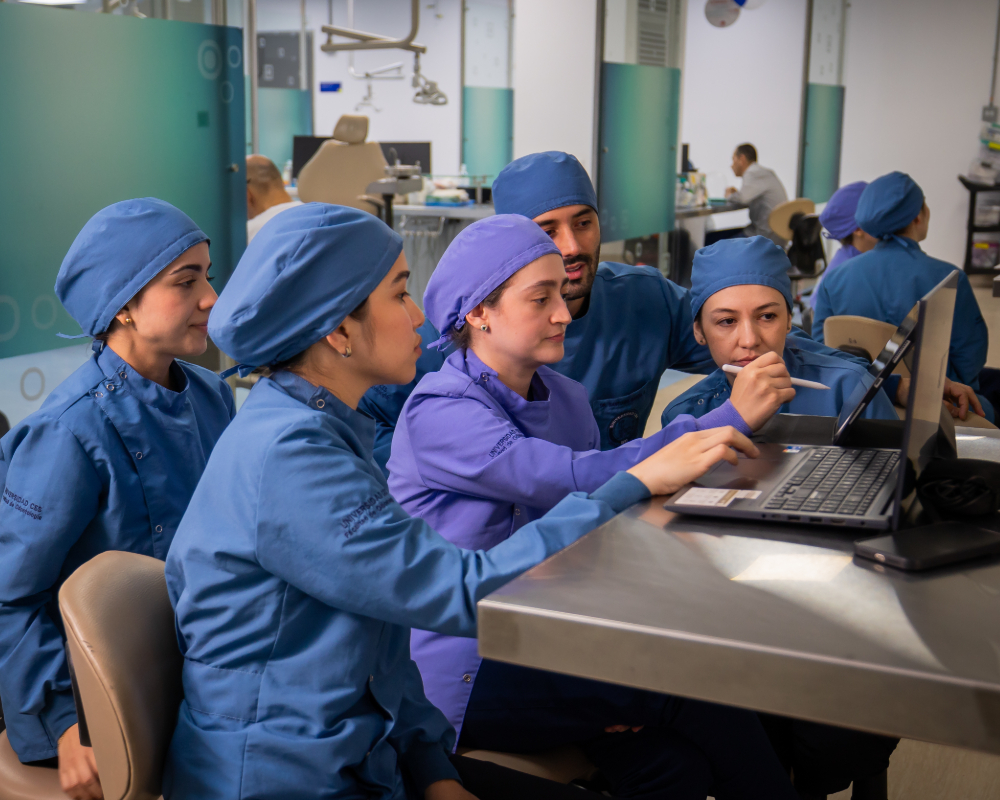 Varias personas con indumentaria clínica están reunidas alrededor de una mesa, observando y trabajando en un computador portátil en un entorno educativo o laboratorio odontológico.