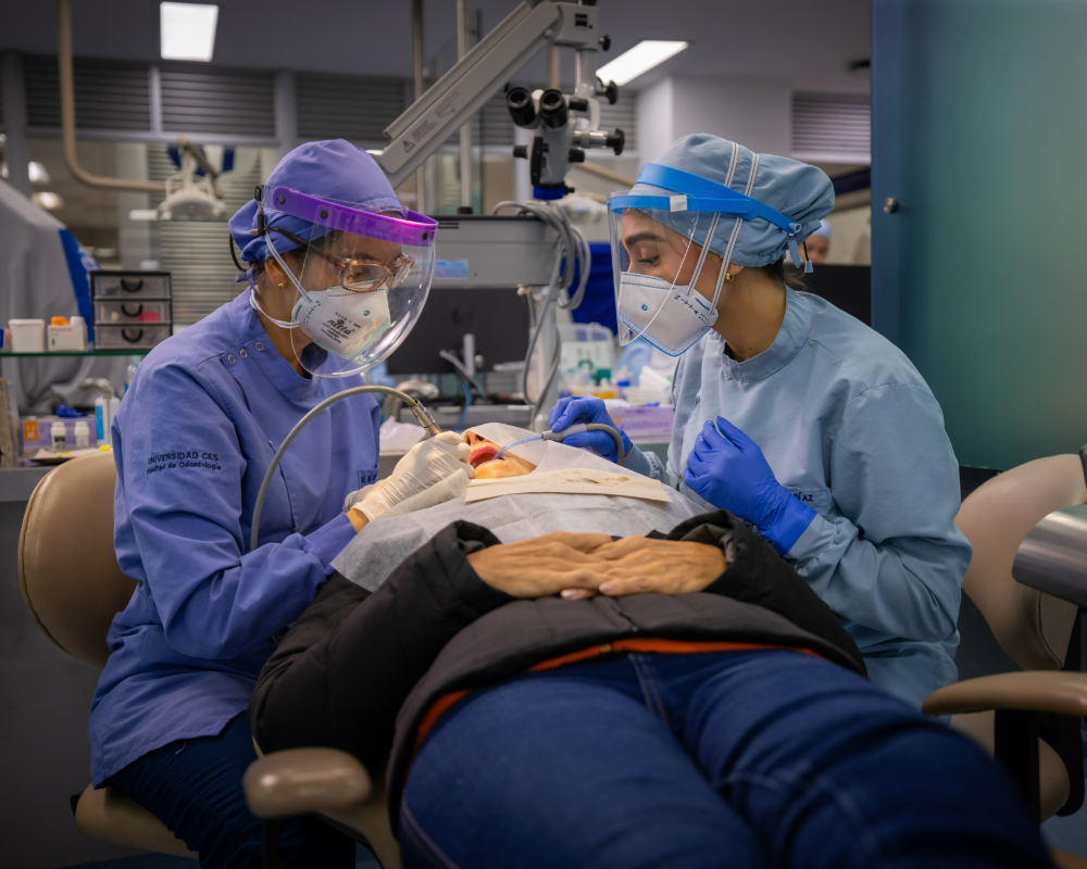Dos profesionales de la salud con uniforme clínico realizan un procedimiento odontológico a una persona recostada en un sillón dental, utilizando instrumentos y equipo especializado.