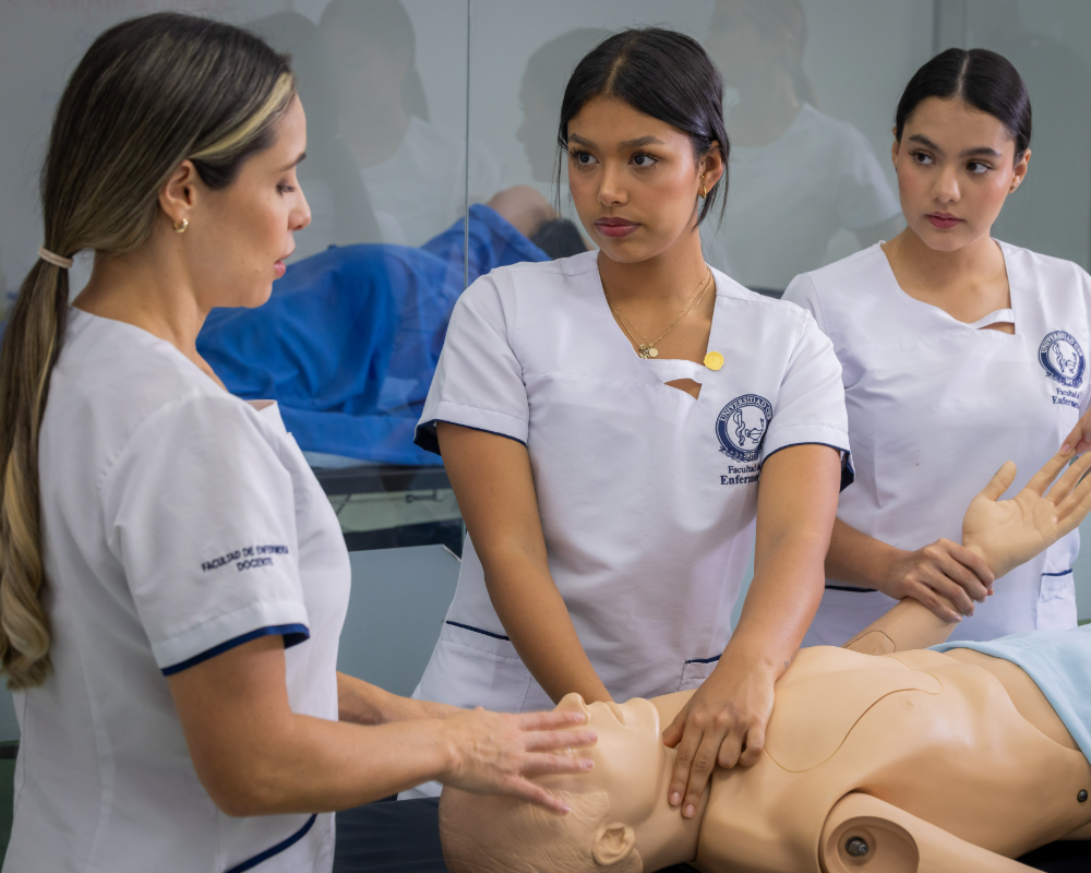 Tres personas con uniformes blancos realizan práctica de reanimación cardiopulmonar (RCP) sobre un simulador de humano en un entorno educativo.