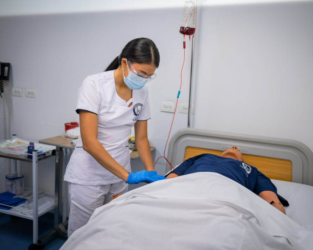Una persona con uniforme clínico coloca guantes y atiende a un paciente recostado en una cama hospitalaria, con equipo médico y una bolsa de suero visible en el entorno.