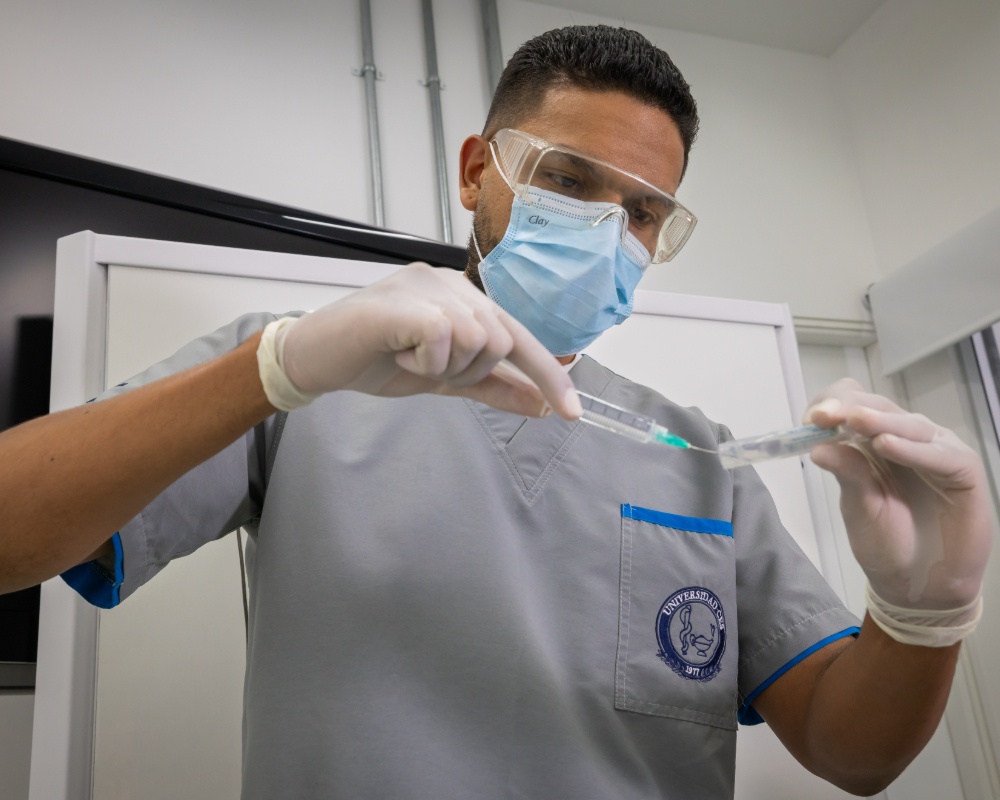 Una persona con uniforme clínico y guantes sostiene y manipula una jeringa en un entorno médico.