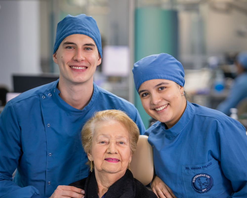 Dos personas con uniforme clínico azul están de pie junto a una persona sentada en un sillón odontológico, en un entorno clínico.