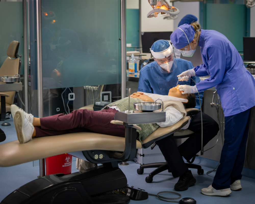 Dos profesionales de la salud realizan un procedimiento odontológico a una persona recostada en un sillón dental dentro de un entorno clínico.