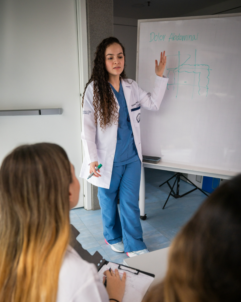 Persona con bata médica explicando un tema en una pizarra frente a otras personas en un entorno educativo.