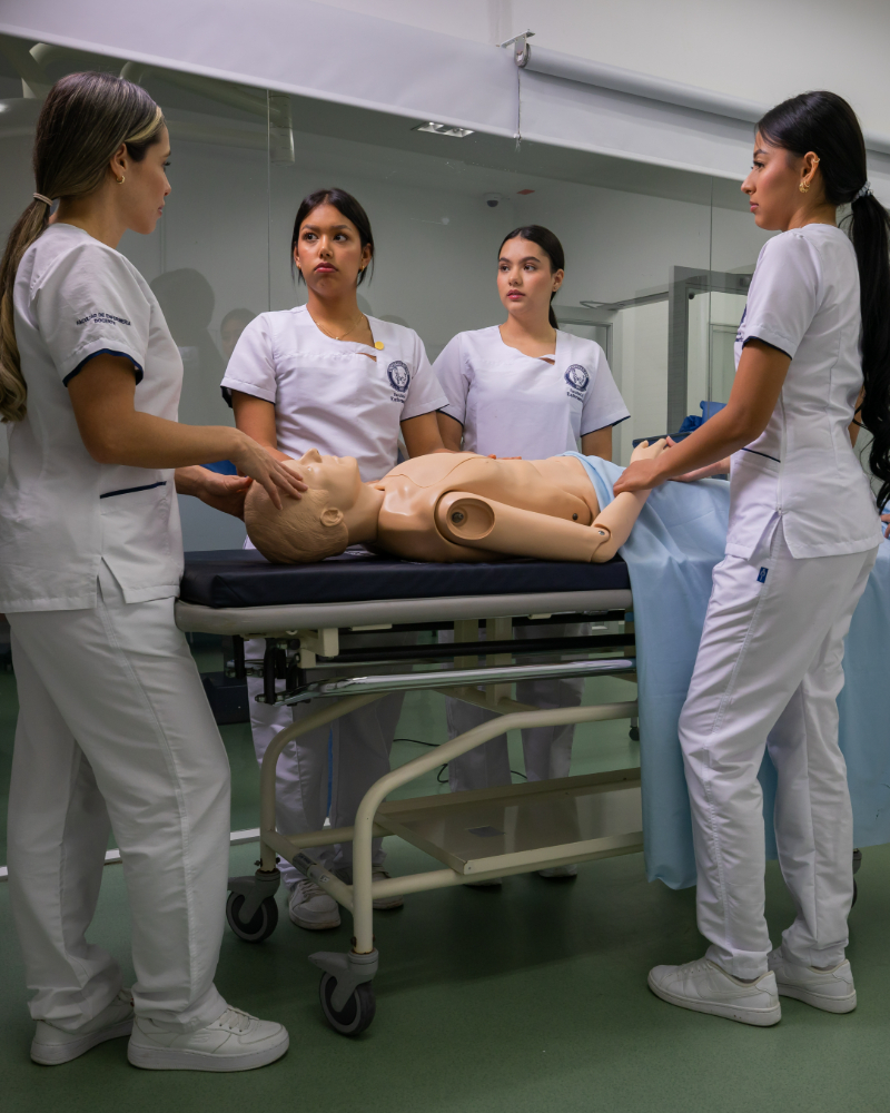 Cuatro personas con uniformes blancos realizan práctica clínica utilizando un simulador humano sobre una camilla en un entorno educativo.