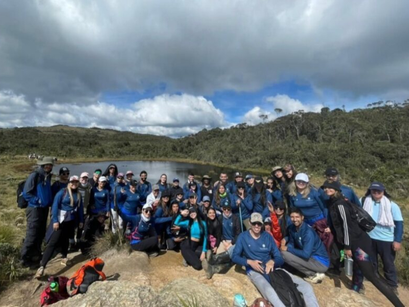 Grupo numeroso de personas vestidas con ropa deportiva azul, reunidas sobre rocas frente a un lago en un paisaje natural con vegetación y cielo parcialmente nublado.