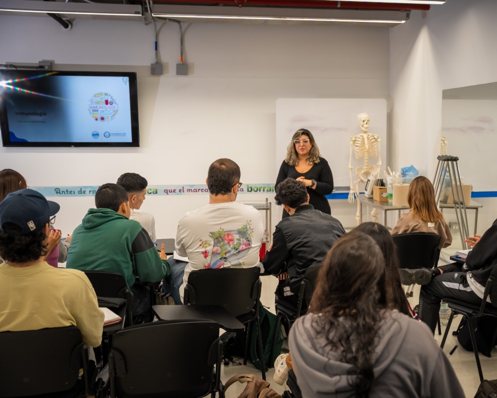 Persona de pie frente a un grupo en un aula, con pantalla encendida, pizarra y un modelo anatómico de esqueleto al fondo.