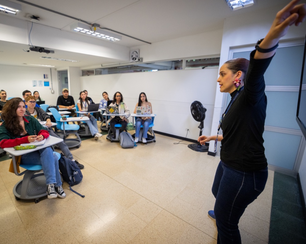 Persona de pie frente a una pantalla en un aula, señalando hacia arriba mientras varias personas están sentadas en escritorios, observando y tomando notas.