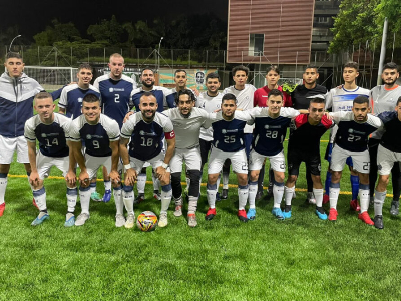 Equipo de fútbol posando en una cancha de césped, con uniformes deportivos blancos y azul oscuro, y un balón en el centro. Al fondo se observan una malla, árboles y un edificio.