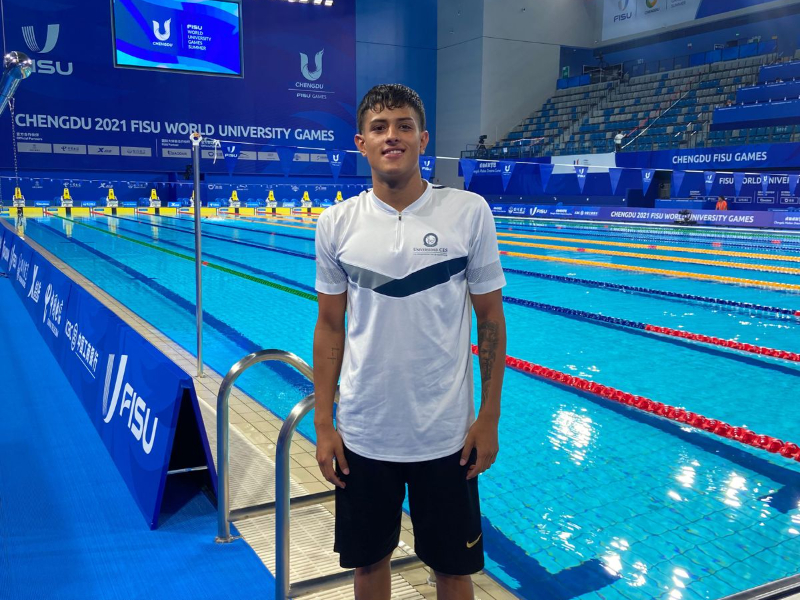 Persona de pie junto a una piscina olímpica en un centro deportivo, con carriles de natación y graderías visibles al fondo. Se observan letreros que indican “Chengdu 2021 FISU World University Games”.