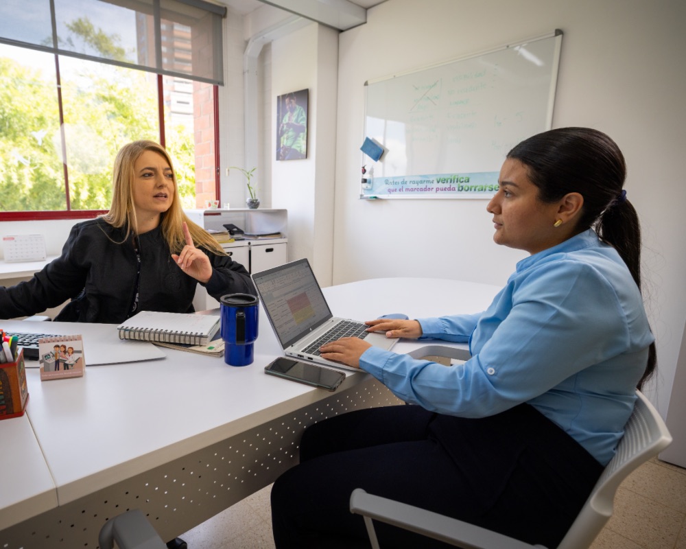Dos personas sentadas en una oficina frente a una mesa blanca; una usa un computador portátil y la otra tiene una libreta y una taza. Al fondo hay una ventana grande y un tablero blanco en la pared.