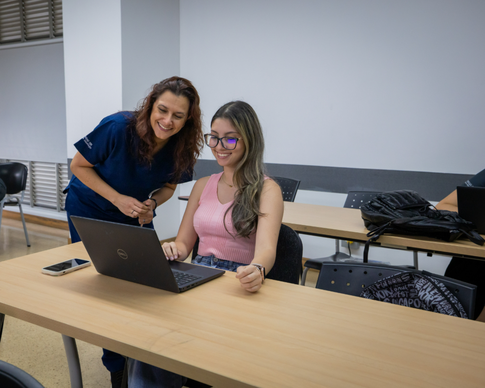 Dos personas en un aula, una sentada frente a un computador portátil y otra de pie junto a ella, sobre mesas largas con sillas.