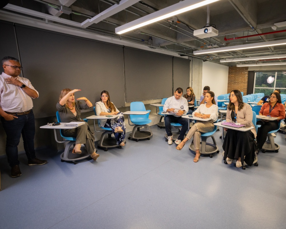 Varias personas sentadas en un aula con sillas azules, escuchando a dos personas.