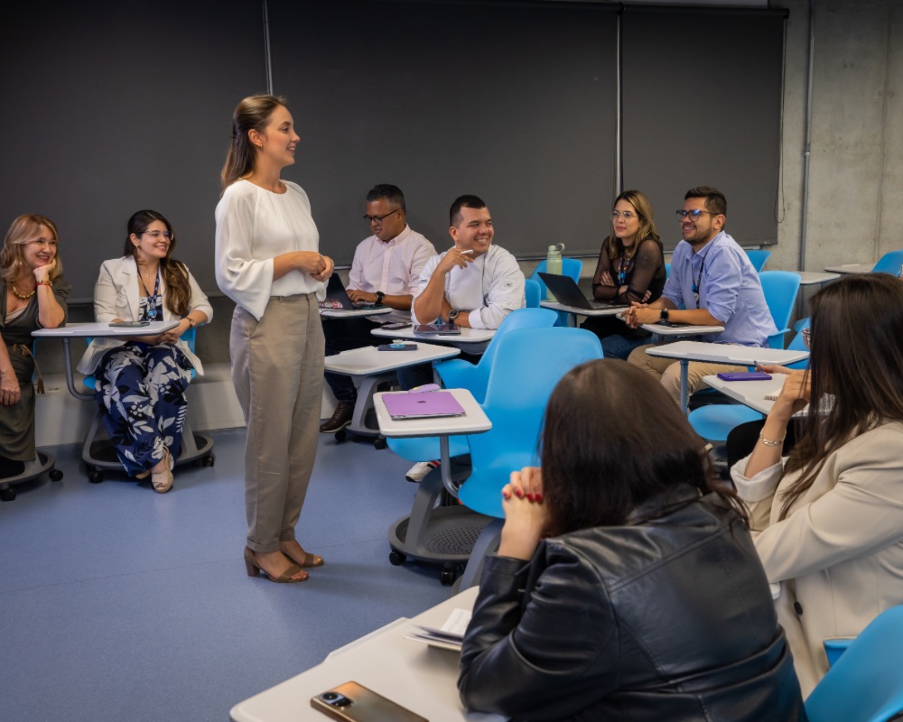 Persona de pie hablando frente a un grupo de personas sentadas en un aula con sillas azules y escritorios individuales.