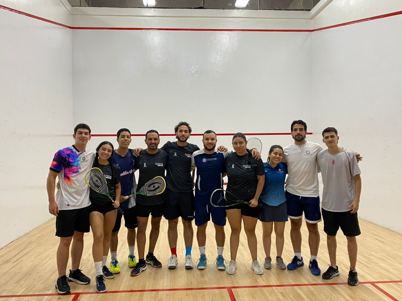 Grupo de personas posando en una cancha de squash, sosteniendo raquetas y vistiendo ropa deportiva, con paredes blancas y líneas rojas características del deporte.