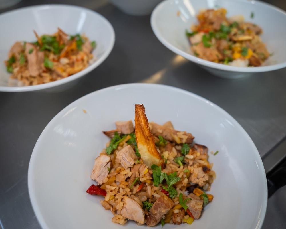 Tres platos hondos contienen arroz mezclado con trozos de pollo, vegetales y hierbas, presentados sobre una superficie metálica.