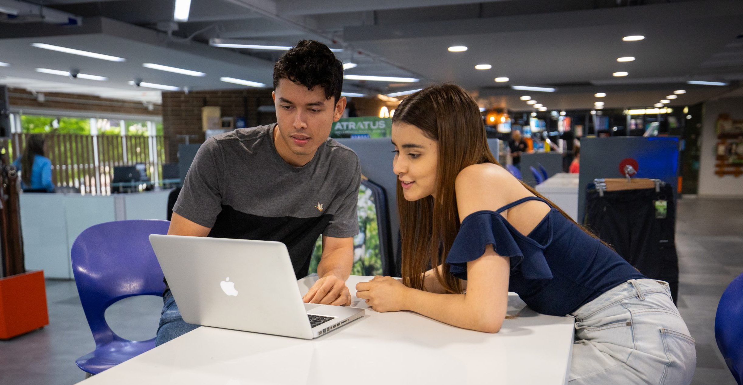 Dos personas sentadas en una mesa alta dentro de un espacio moderno; sobre la mesa hay un computador portátil abierto y ambos están mirando la pantalla. Al fondo se observan sillas, estanterías y elementos decorativos.