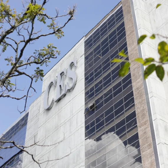 Fachada de un edificio institucional con el nombre ‘CES’, con superficie de vidrio y concreto, vista en ángulo bajo junto a ramas de árboles y cielo despejado.