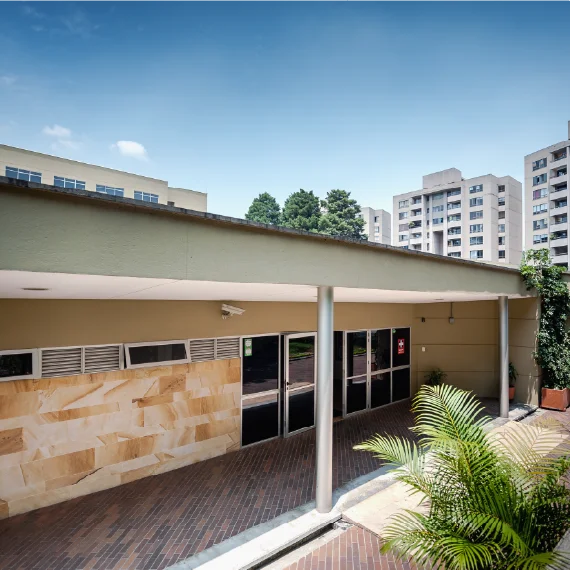 Entrada de una edificación institucional con puertas de vidrio y pared en tonos beige, cubierta por un techo con columnas, en un entorno urbano con edificios y vegetación.