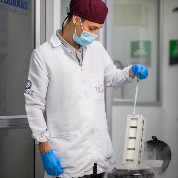 Fotografía de una persona con bata de laboratorio, mascarilla y guantes azules manipulando un instrumento de medición sobre un equipo que emite vapor. La escena se desarrolla en un laboratorio y representa actividades de investigación científica y trabajo experimental.